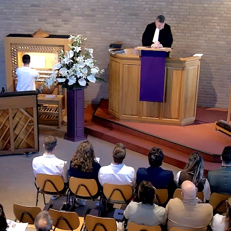Screendump van de livestream met predikant op kansel, organist en een paar mensen op de voorste rij in de kerk